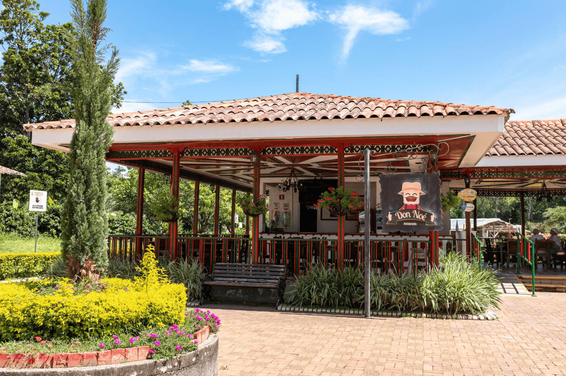 Restaurante al aire libre con techo de tejas, rodeado de vegetación y flores, con un cartel de madera con un chef de dibujos animados y el nombre "Don Noel", que forma parte de La Familia Noé. El cielo azul y los árboles crean un telón de fondo encantador.