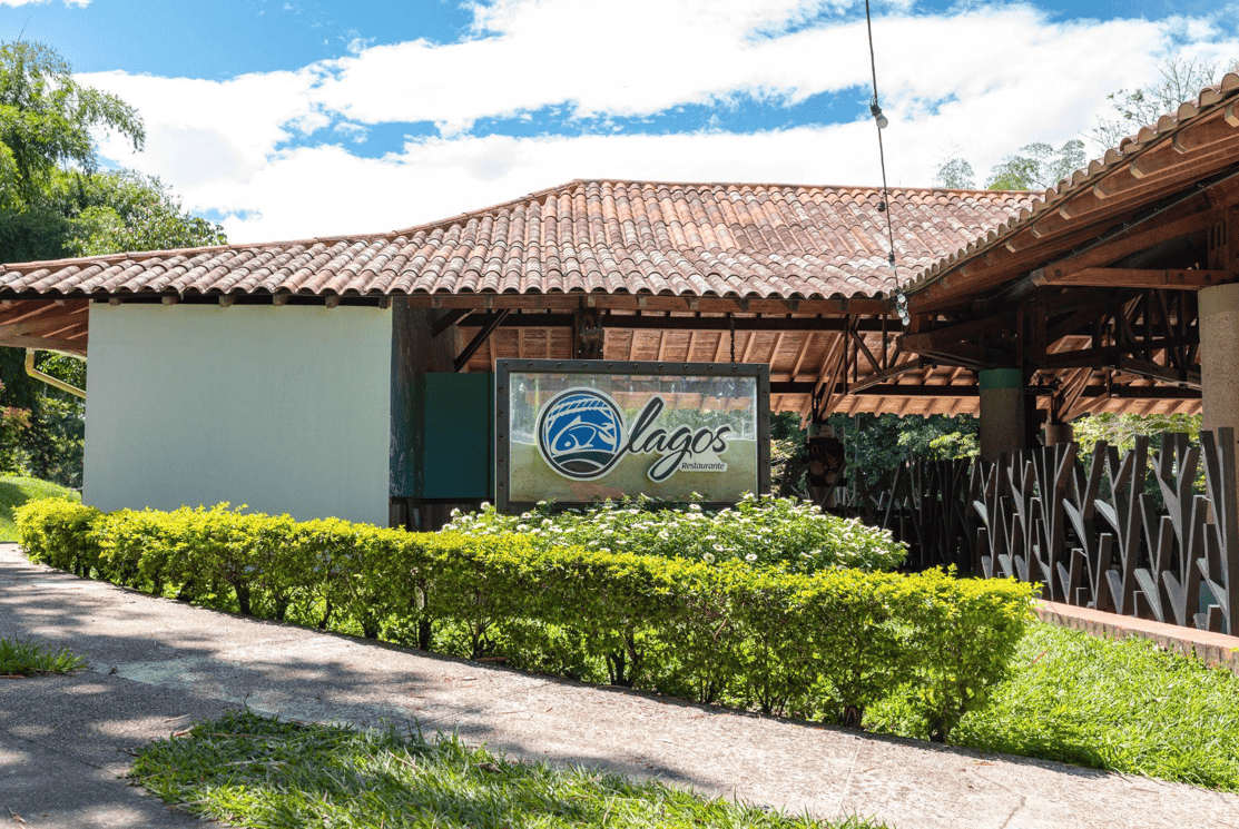 Vista exterior de un edificio rústico con tejado de tejas, rodeado de vegetación. Delante hay un cartel en el que se lee Lagos con el logotipo de un pez azul, lo que indica que puede tratarse de un restaurante o un área recreativa.