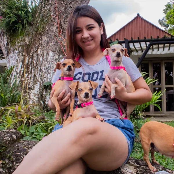 Una mujer sentada al aire libre, sonriendo y sujetando a tres pequeños perros de color canela con arneses rosas. Otro perro está a su lado. Rodeada por un gran árbol y una casa, disfruta de un momento de paz en Mundo Rural con sus compañeros de Mundo Animal.