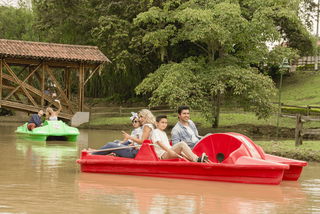 La gente disfruta de un relajante paseo en bote, montada en botes de pedales rojos y verdes a lo largo de un río tranquilo y arbolado. Un puente de madera cruza el agua al fondo mientras todos disfrutan de su apacible paseo en este día nublado.