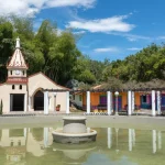 Una pequeña capilla, conocida como Capilla Pereira, con una torre de reloj, se alza junto a coloridos edificios y una fuente circular en una plaza, rodeada de exuberante vegetación bajo un cielo azul brillante con nubes dispersas.