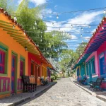 Cerca de Capilla Pereira Antigua se extiende una calle adoquinada bordeada de casas de colores en naranja, verde, rosa y azul brillantes, con luces de cuerda por encima y bancos a los lados bajo un cielo soleado y azul.