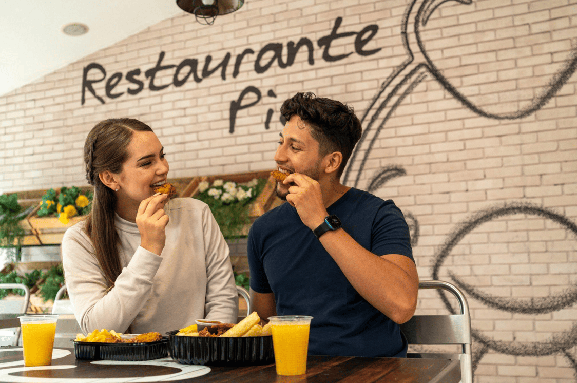 Una mujer y un hombre sonrientes se sientan a la mesa de un restaurante y comparten una comida a base de pollo frito, patatas fritas y zumo de naranja. El nombre del restaurante está pintado en una pared de ladrillo blanco detrás de ellos.