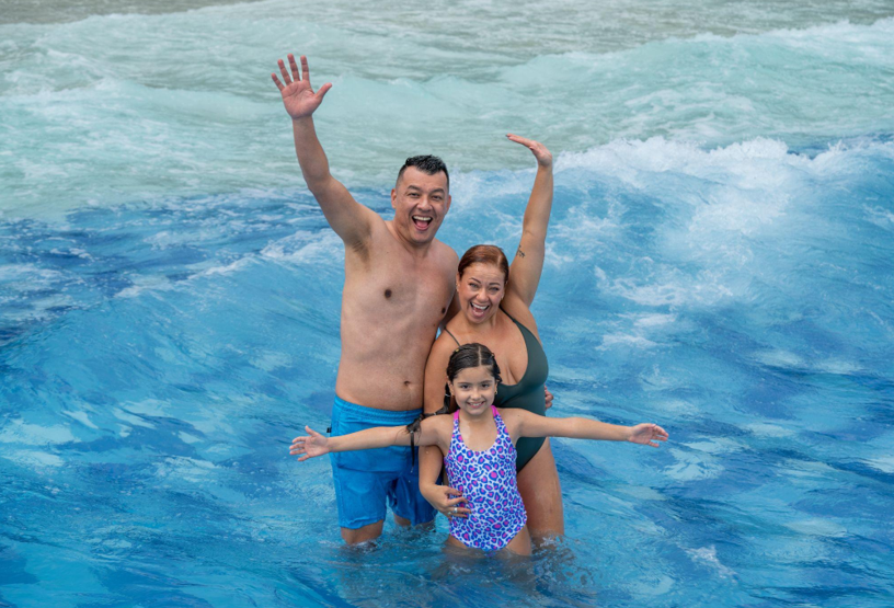 Una sonriente familia de tres miembros, dos adultos y un niño en bañador, permanecen juntos en una piscina de olas con los brazos en alto, disfrutando del agua y del chapoteo de las olas a su alrededor.