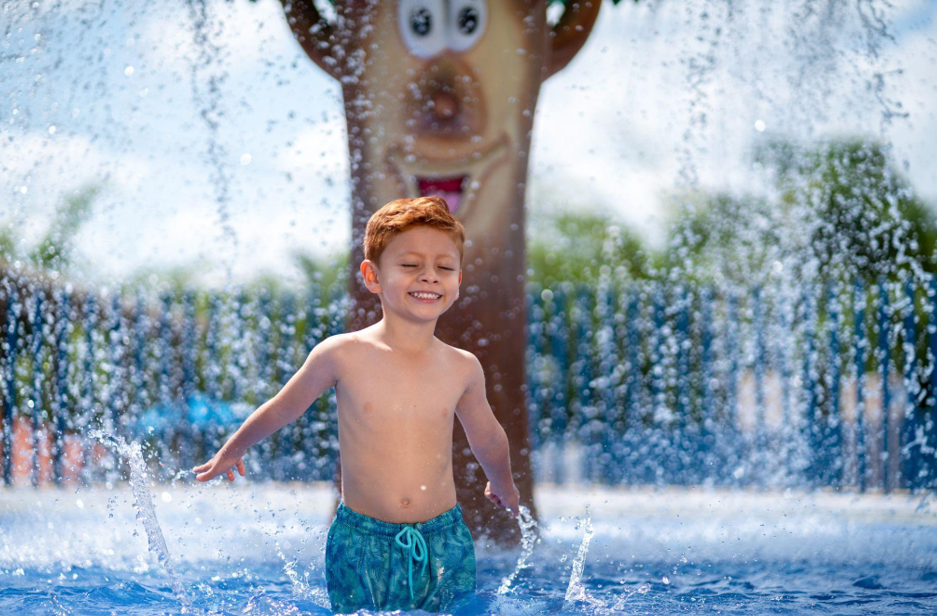Un niño con bañador azul sonríe con los ojos cerrados mientras juega en las fuentes de agua de un parque acuático, cerca de una estructura de juegos con forma de oso y una colorida piscina honguito al fondo.