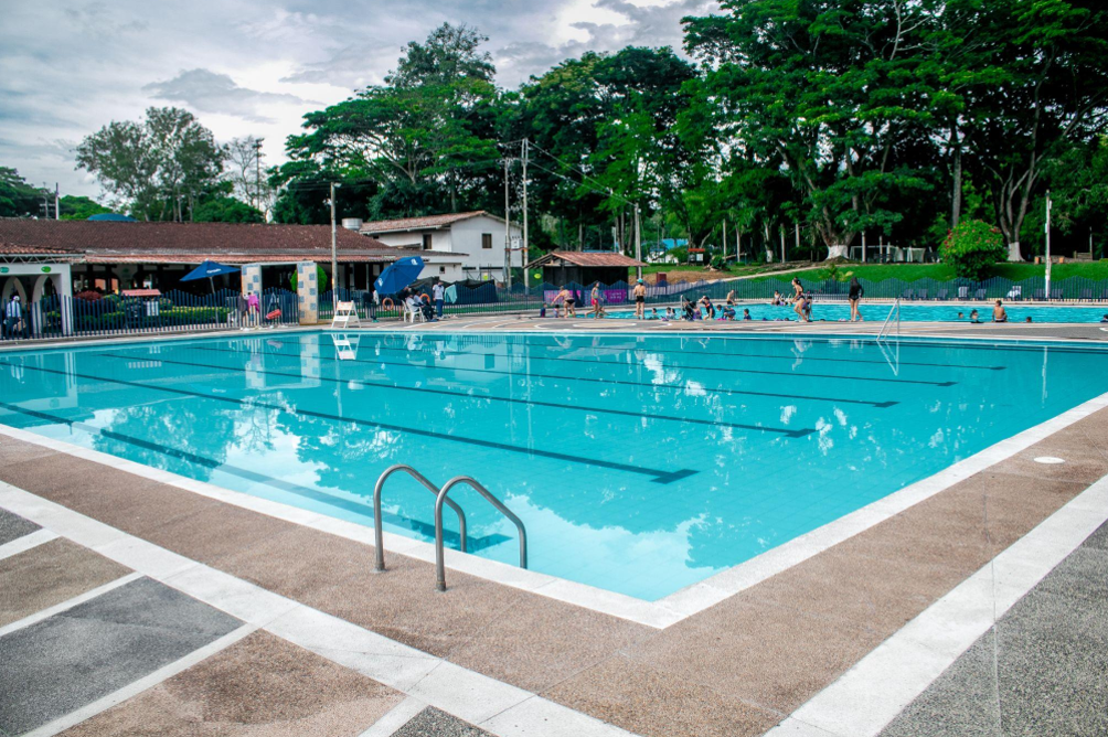 Una gran piscina al aire libre con agua azul clara, rodeada de una cubierta de azulejos. La gente se relaja alrededor de las piscinas gemelas, con árboles y edificios de fondo bajo un cielo nublado.