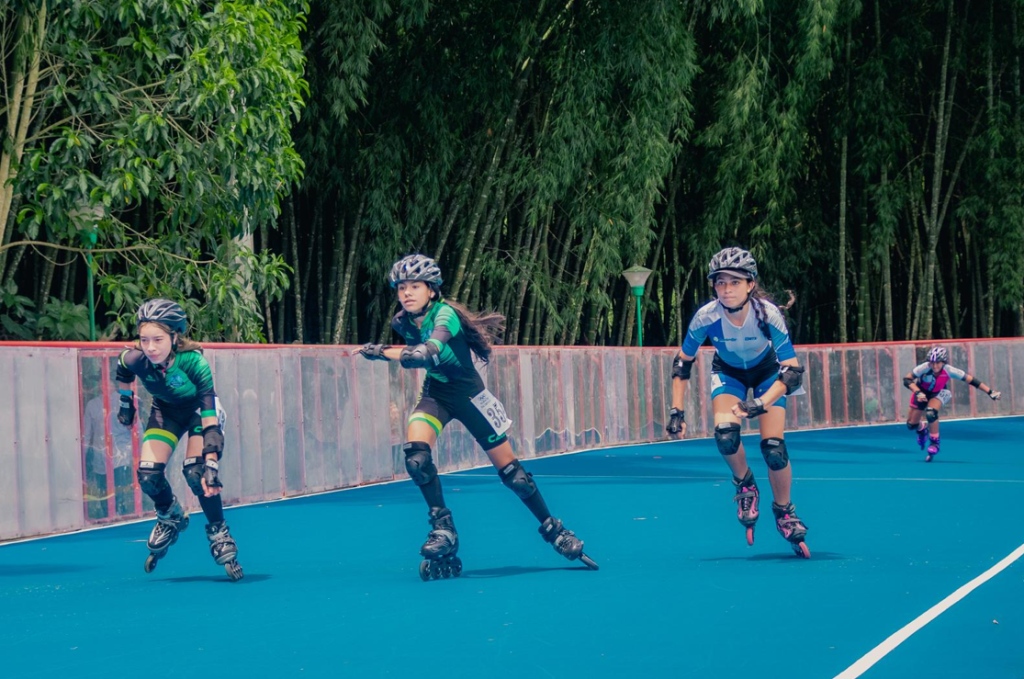Cuatro jóvenes atletas, con cascos y protecciones, corren en patines alrededor de una pista de patinaje azul rodeada de frondosos árboles verdes y una barrera transparente. Tres compiten de cerca en cabeza, mientras que un patinador va detrás.