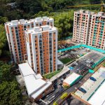 Vista aérea de dos modernos edificios de apartamentos rodeados de árboles, con un aparcamiento y coches en medio, y una grúa de construcción cerca de un tercer edificio en construcción.