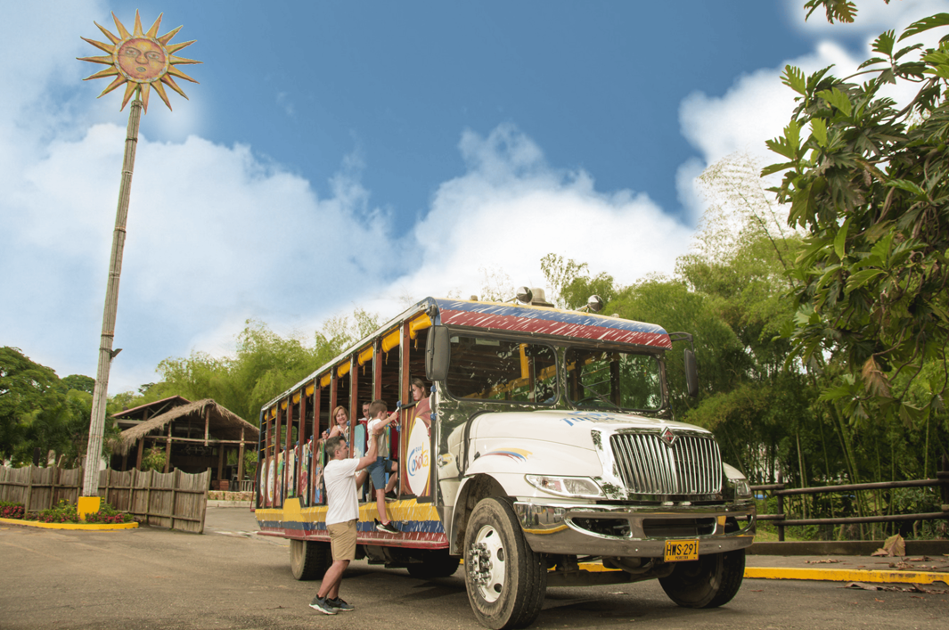 Un colorido autobús abierto para un recorrido en chiva con pasajeros está aparcado cerca de vegetación tropical. Un hombre está de pie junto al autobús, y un poste alto con una cara de sol decorativa remata la escena bajo un cielo azul brillante con nubes dispersas.