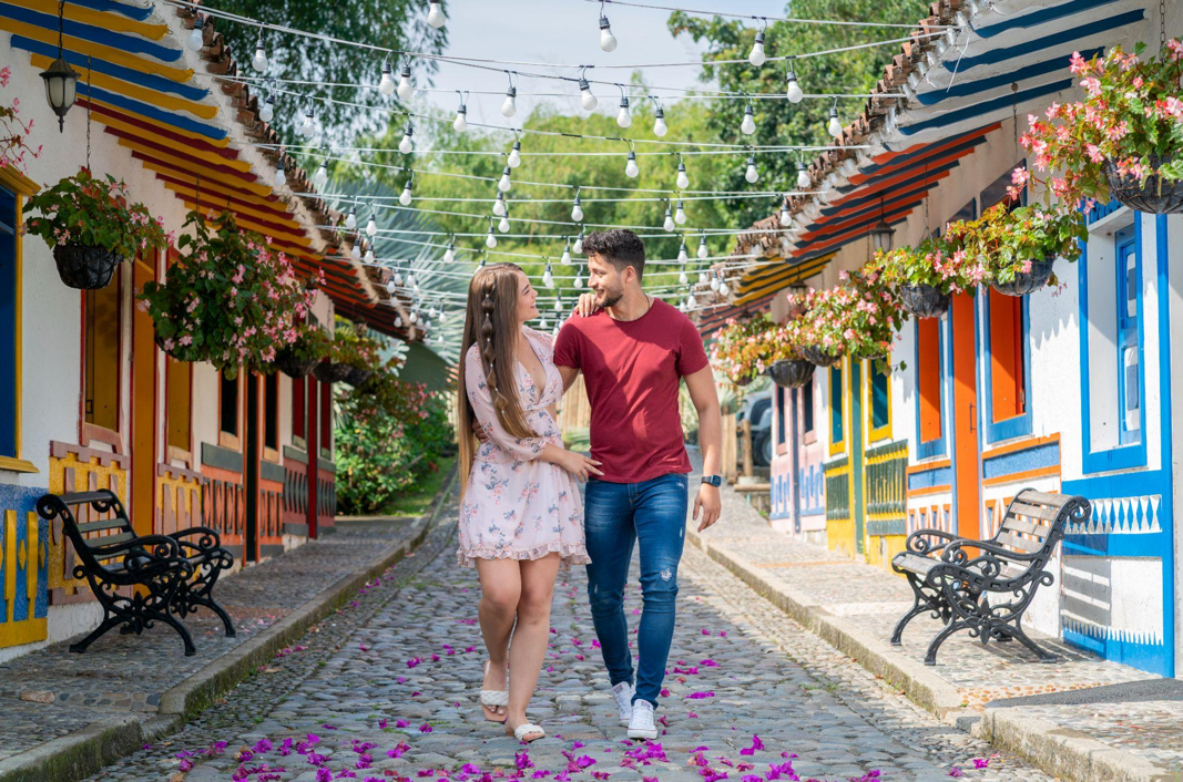 Una pareja camina cogida de la mano por una calle adoquinada y sembrada de pétalos de flores en Pereira Antigua, bordeada de casas de colores, bancos y cestas de flores colgantes bajo cuerdas de luces, una encantadora réplica del vibrante pasado de Pereira.
