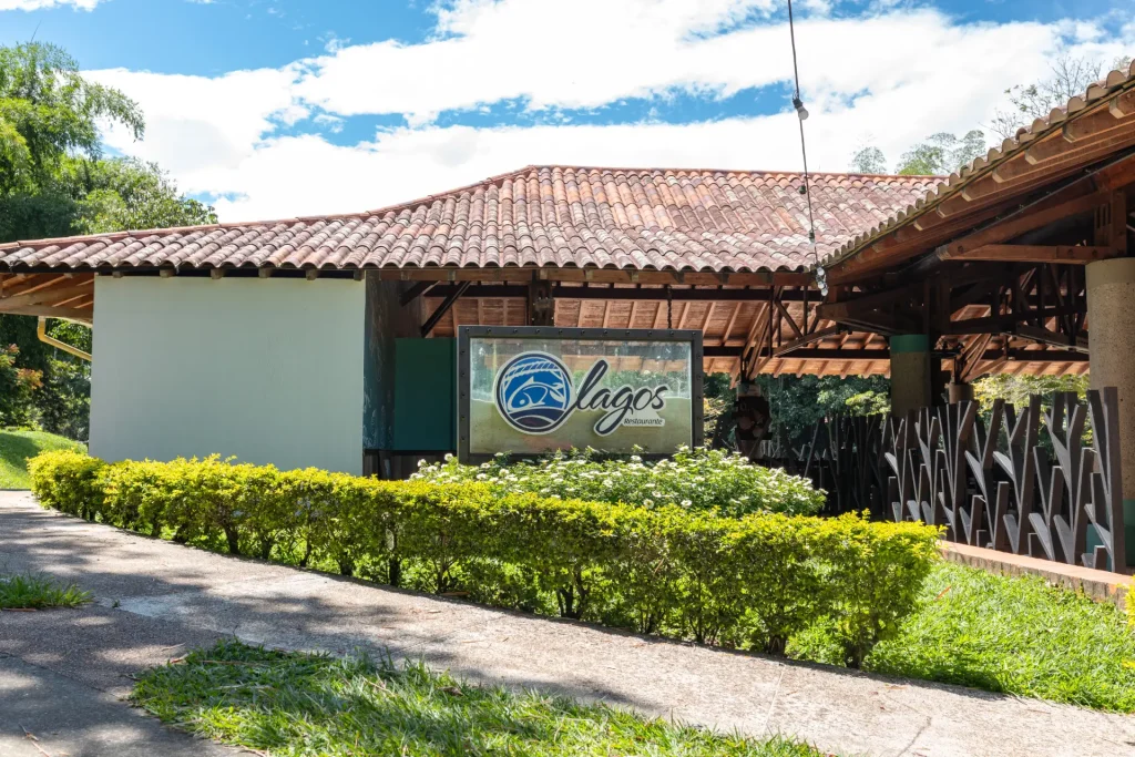 Entrada del restaurante Lagos con un cartel con el logotipo del restaurante, rodeado de vegetación, arbustos y un edificio con tejado de tejas al fondo, bajo un cielo parcialmente nublado.