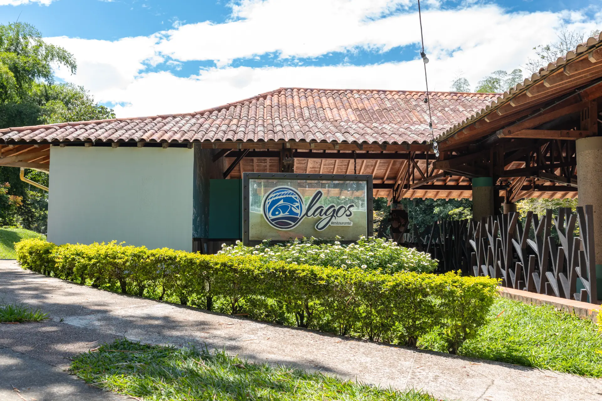 Entrada del restaurante Lagos con un cartel con el logotipo del restaurante, rodeado de vegetación, arbustos y un edificio con tejado de tejas al fondo, bajo un cielo parcialmente nublado.