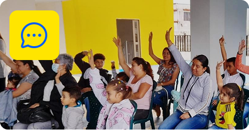 Un grupo de adultos y niños sentados en sillas en el interior, algunos levantando la mano, como si compartieran ideas sobre construir un hogar. El fondo muestra una pared y una puerta de color amarillo brillante, con un icono de chat amarillo y líneas azules superpuestas en la esquina superior izquierda.