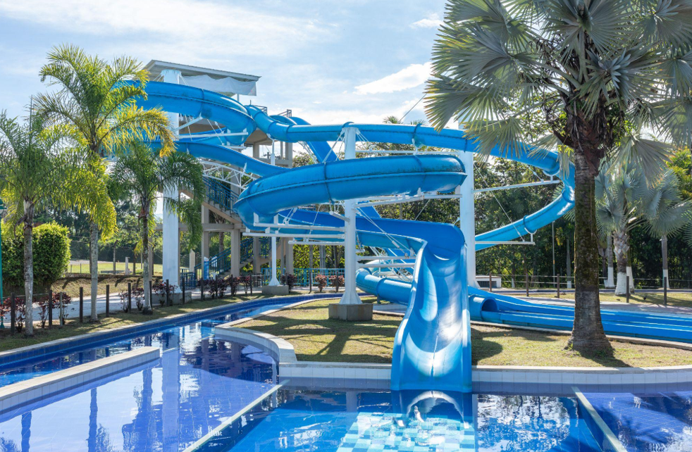 Un gran tobogán azul se curva sobre una piscina en un parque acuático al aire libre, rodeado de palmeras y vegetación en un día soleado.