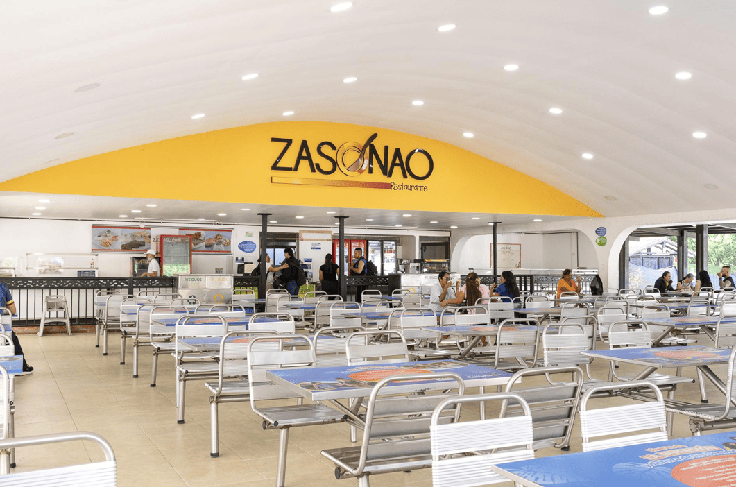 Una cafetería espaciosa y bien iluminada, con mesas y sillas metálicas vacías, algunas personas sentadas al fondo y una pared amarilla con el rótulo Zasonao restaurante sobre el mostrador de servicio.