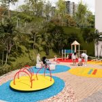 Niños y adultos disfrutan de una colorida zona de juegos al aire libre en el Parque Arboleda, con un tiovivo, estructuras para trepar, un camino para jugar a la rayuela y un tobogán, todo ello rodeado de árboles y vegetación cerca de altos edificios.