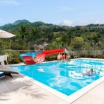 En el Parque Arboleda, una soleada piscina al aire libre muestra a niños jugando en un tobogán rojo y en el agua, mientras una mujer se relaja en una tumbona y otra se sienta en una mesa bajo una sombrilla, con exuberantes colinas verdes de fondo.