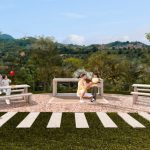 Dos bancos enfrentados en un patio pavimentado del parque Arboleda. Una mujer y un niño juegan con un avión de juguete cerca, mientras otra mujer y otro niño se sientan juntos sosteniendo un globo rojo. Árboles y montañas forman el pintoresco fondo.