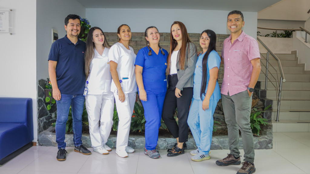 Un grupo de siete personas, entre ellas profesionales sanitarios uniformados y dos hombres con ropa informal, permanecen juntos en el interior, sonriendo a la cámara y representando con orgullo a Nosotros UID.