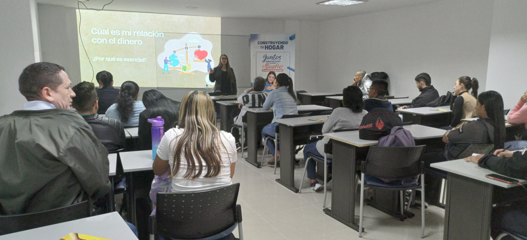 Una mujer está de pie delante de un aula dando una presentación sobre las relaciones con el dinero a adultos sentados. Una diapositiva detrás de ella dice: ¿Cuál es mi relación con el dinero? ¿Por qué es esencial? -clave para construir un hogar estable.