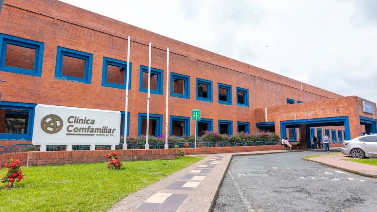 Un edificio de ladrillo rojo con ventanas de marco azul alberga la Clínica Comfamiliar, que ofrece pólizas y medicina prepagada. Delante se ven banderas, flores y un camino de entrada curvado, con un par de personas cerca de la entrada en un día nublado.