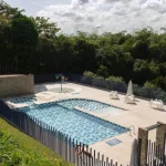 La piscina exterior del Hotel Chapola está rodeada por una valla y árboles, y cuenta con una zona poco profunda para niños, tumbonas y sombrillas en la cubierta de baldosas. La piscina está casi vacía bajo un cielo parcialmente nublado.