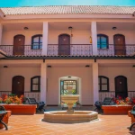 Patio abierto del Hotel Chapola con una fuente central de piedra, rodeada de bancos y parterres. Edificio de dos plantas con puertas de madera arqueadas, barandillas de hierro y tejado de tejas bajo un cielo azul brillante.