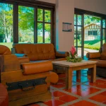 La acogedora zona de estar del Hotel Chapola cuenta con sillones de cuero marrón y un sofá a juego alrededor de una mesa de centro de madera con un arreglo floral. Los grandes ventanales dejan ver un exuberante jardín exterior y un pequeño edificio.