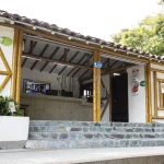 Un edificio rústico con entramado de bambú, tejado de tejas y escalones de piedra que conducen a una entrada abierta. En el interior del Hotel Chapola hay mostradores, carteles informativos y plantas verdes, con árboles visibles al fondo.