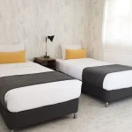Un dormitorio luminoso y moderno del Hotel Chapola cuenta con dos camas individuales con ropa de cama blanca, mantas grises y cojines amarillos. Junto a cada cama hay mesillas de noche de madera y lámparas negras, y por la ventana entra la luz del sol.