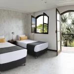 Una luminosa habitación del Hotel Chapola con dos camas individuales, almohadas amarillas, un televisor montado en la pared y puertas abiertas que dan a un pequeño balcón con vistas a las palmeras y a la exuberante vegetación del exterior.