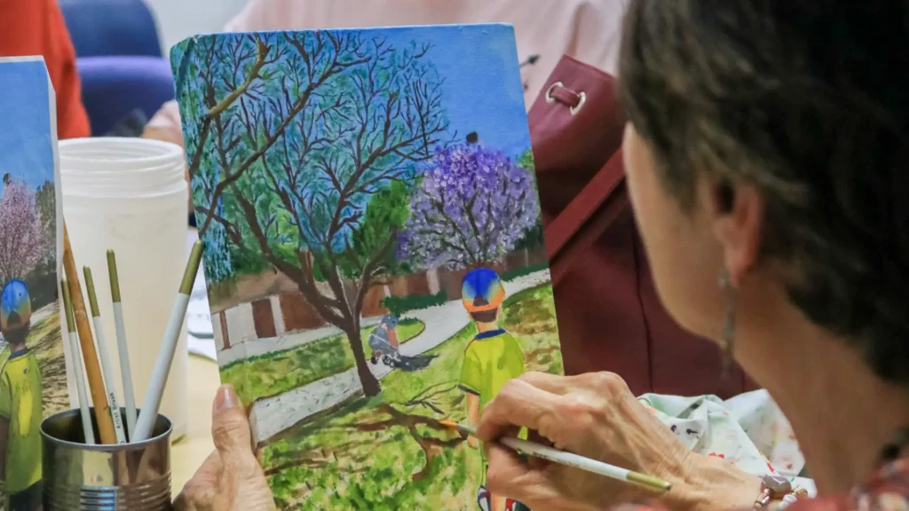 Una persona sostiene y pinta un colorido cuadro de una escena de parque con árboles y dos niños, uno de ellos con camiseta amarilla y gorra azul, mientras está sentada a una mesa con pinceles y material de arte cerca.