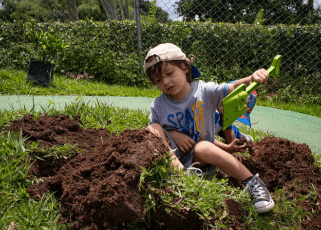 Un niño pequeño con gorra y zapatillas de deporte está sentado sobre la hierba, cavando en la tierra con una pala verde de juguete. Manos a la granja, exploran la tierra con curiosidad, un montículo de tierra ante ellos y una frondosa vegetación más allá de la valla.