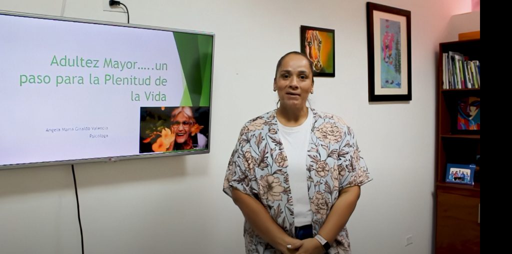 Una mujer se encuentra en un despacho junto a una pantalla que muestra una presentación titulada Adultez Mayor... un paso para la Plenitud de la Vida, que nos recuerda que envejecer es construir sueños. Arte y libros decoran la pared y la estantería detrás de ella.