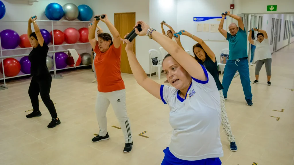 Un grupo de adultos en ropa de ejercicio se estiran con los brazos en alto, sujetando pesas de mano, en una sala de fitness muy iluminada con pelotas de ejercicio en estanterías al fondo.
