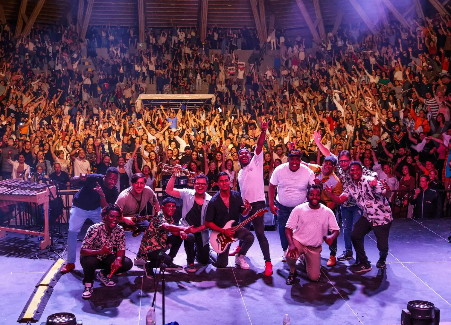 Una banda posa junta en la parte delantera de un escenario, sonriendo y sujetando instrumentos, mientras un público entusiasta anima de fondo en este animado concierto en interior que celebra Afiliación Independientes.