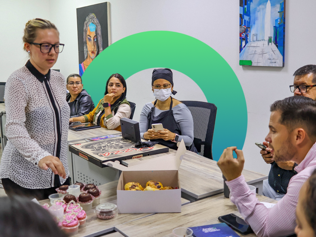 Una mujer junto a una mesa con pizza, magdalenas y pasteles durante un evento de la Ruta de emprendimiento; sus colegas se sientan alrededor con portátiles y bebidas. Una persona lleva una máscara y un gorro de cocinero; coloridas obras de arte alegran las paredes de la oficina.