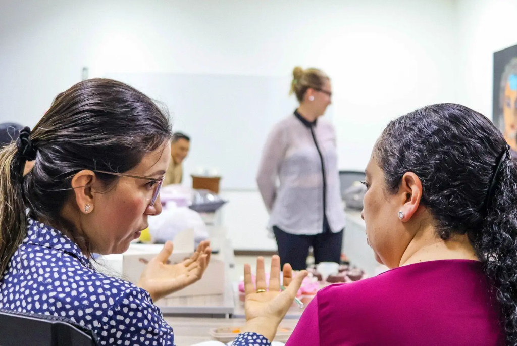 Dos mujeres se sientan y hablan de su ruta de emprendimiento, una gesticulando con las manos, mientras otras dos personas permanecen borrosas al fondo en un interior muy iluminado.