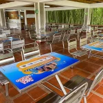 Cafetería al aire libre en la Zona de Eventos, con mesas y sillas metálicas vacías sobre un suelo de baldosas rojas. Algunas mesas exhiben coloridos carteles promocionales. El bambú verde de Los Helechos se ve a través de las paredes abiertas.