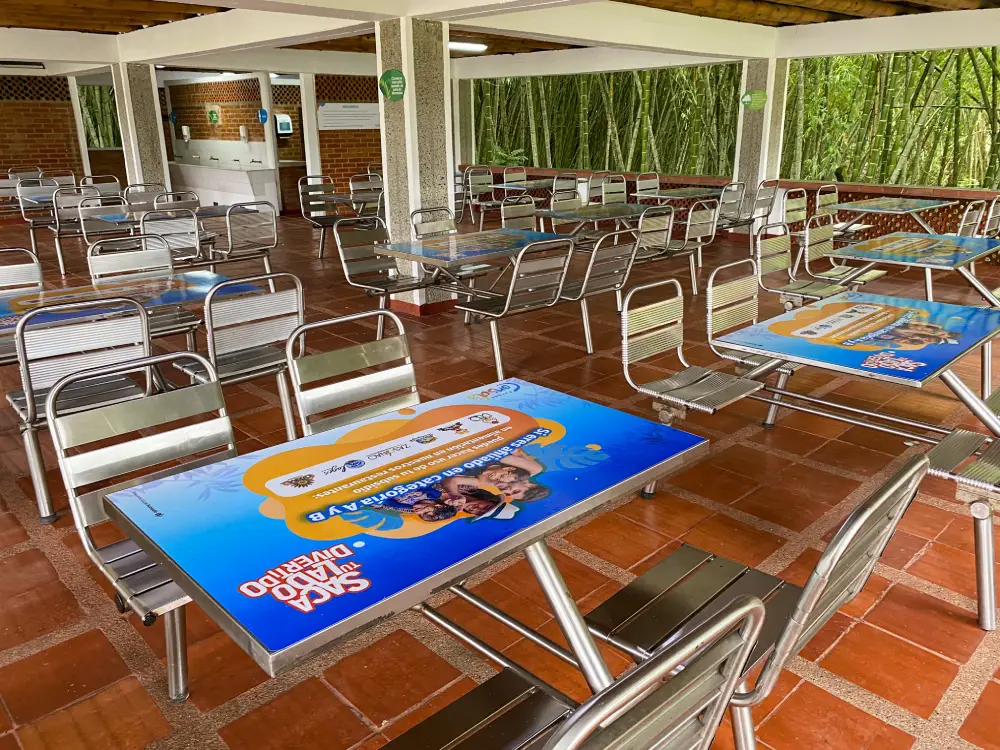 Cafetería al aire libre en la Zona de Eventos, con mesas y sillas metálicas vacías sobre un suelo de baldosas rojas. Algunas mesas exhiben coloridos carteles promocionales. El bambú verde de Los Helechos se ve a través de las paredes abiertas.