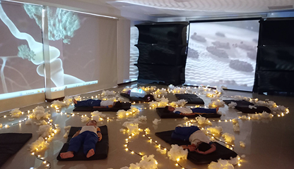 La gente está tumbada en colchonetas en una sala poco iluminada, decorada con cuerdas de luz y objetos blancos en forma de nube. En las paredes se proyectan grandes imágenes científicas, creando una atmósfera envolvente y tranquilizadora que fomenta la educación inclusiva.