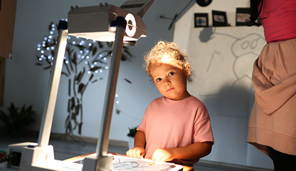 Un niño pequeño con una camisa rosa está de pie bajo la luz de un retroproyector, mirando pensativo hacia arriba. Dibujos y adornos reflejan el enfoque Reggio y los lenguajes de los niños en el fondo suavemente iluminado.