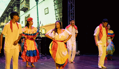 Cinco personas ataviadas con coloridos trajes tradicionales interpretan una danza folclórica en el escenario, celebrando el arte y la cultura. Los bailarines visten trajes blancos con toques rojos y amarillos; dos mujeres llevan faldas con volantes y tocados de flores.
