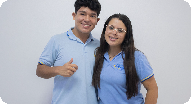Dos personas vestidas con camisas de cuello azul claro permanecen juntas, sonriendo a la cámara en un ambiente de modalidad presencial. La persona de la izquierda levanta el pulgar, mientras que la de la derecha lleva gafas. El fondo es blanco liso.