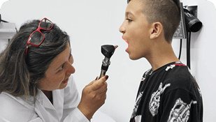 Un médico con bata blanca examina la garganta de un niño con un instrumento médico, buscando signos de enfermedades congénitas mientras el niño permanece sentado con la boca abierta.