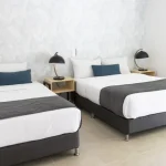 Un dormitorio luminoso y moderno en Cabañas Consotá cuenta con dos camas con ropa de cama blanca, mantas grises y almohadas azules. Dos lámparas negras descansan sobre mesitas de noche de madera entre las camas, y a la derecha hay una ventana con una persiana a rayas.