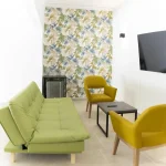 El moderno salón de Cabañas Consotá cuenta con un futón verde, dos sillas amarillo mostaza, una mesa de centro rectangular de madera, un televisor montado en la pared y una pared de acento con motivos de hojas tropicales.