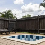 Una pequeña piscina cuadrada con azulejos azules y blancos está situada en una terraza de madera junto a una tumbona negra en Cabañas Consotá. La zona está delimitada por una alta valla de madera oscura y rodeada de palmeras bajo un cielo parcialmente nublado.