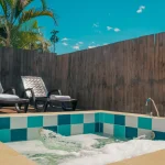 Una pequeña bañera de hidromasaje al aire libre con agua burbujeante se encuentra junto a dos tumbonas acolchadas y palmeras en Cabañas Consotá, todo ello rodeado por una valla de privacidad de madera bajo un cielo azul claro.