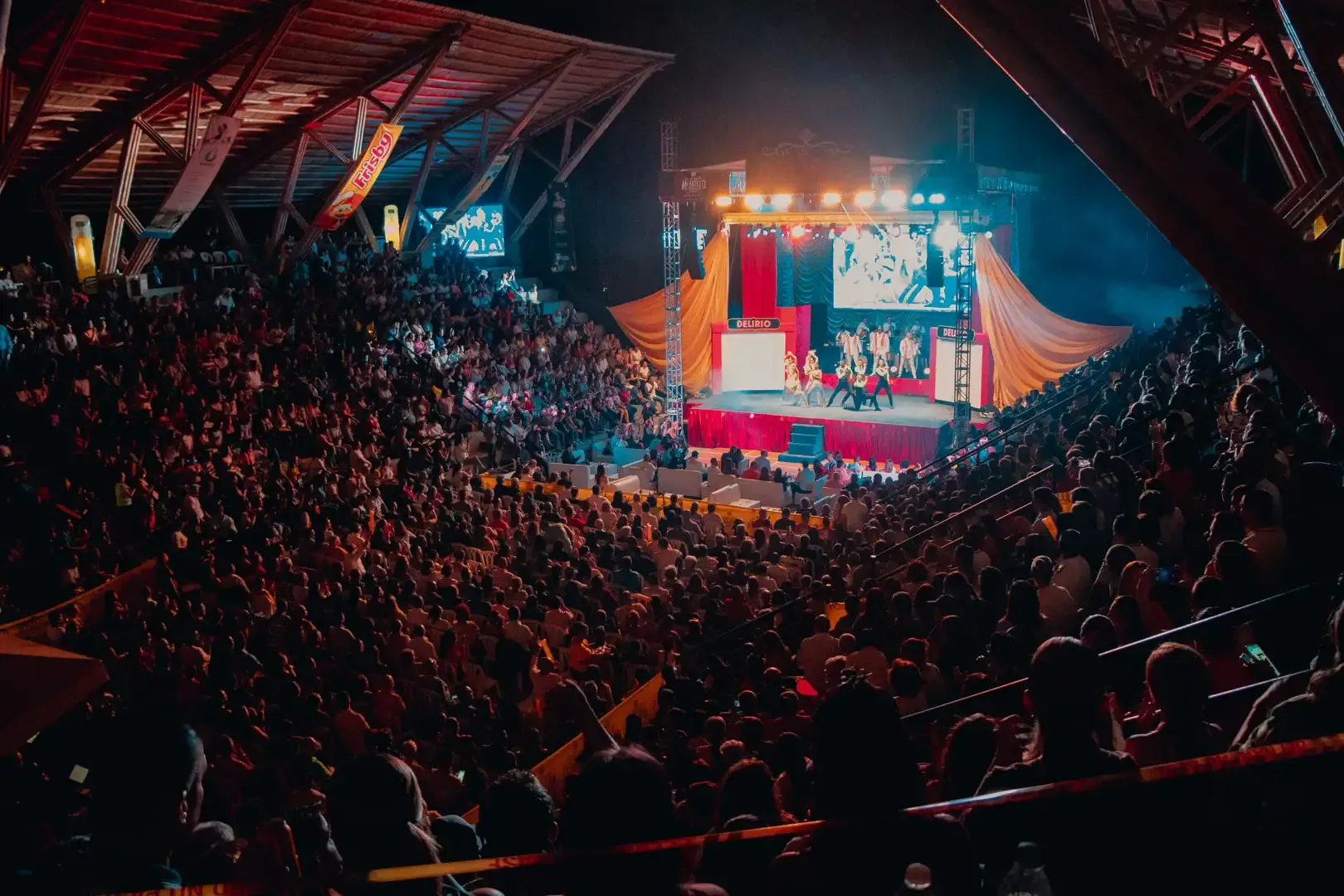 Una gran multitud llena el Coliseo Guaduales, observando un escenario brillantemente iluminado con telones rojos y artistas. El público se sienta en filas escalonadas, mientras las pantallas muestran la acción en el escenario.